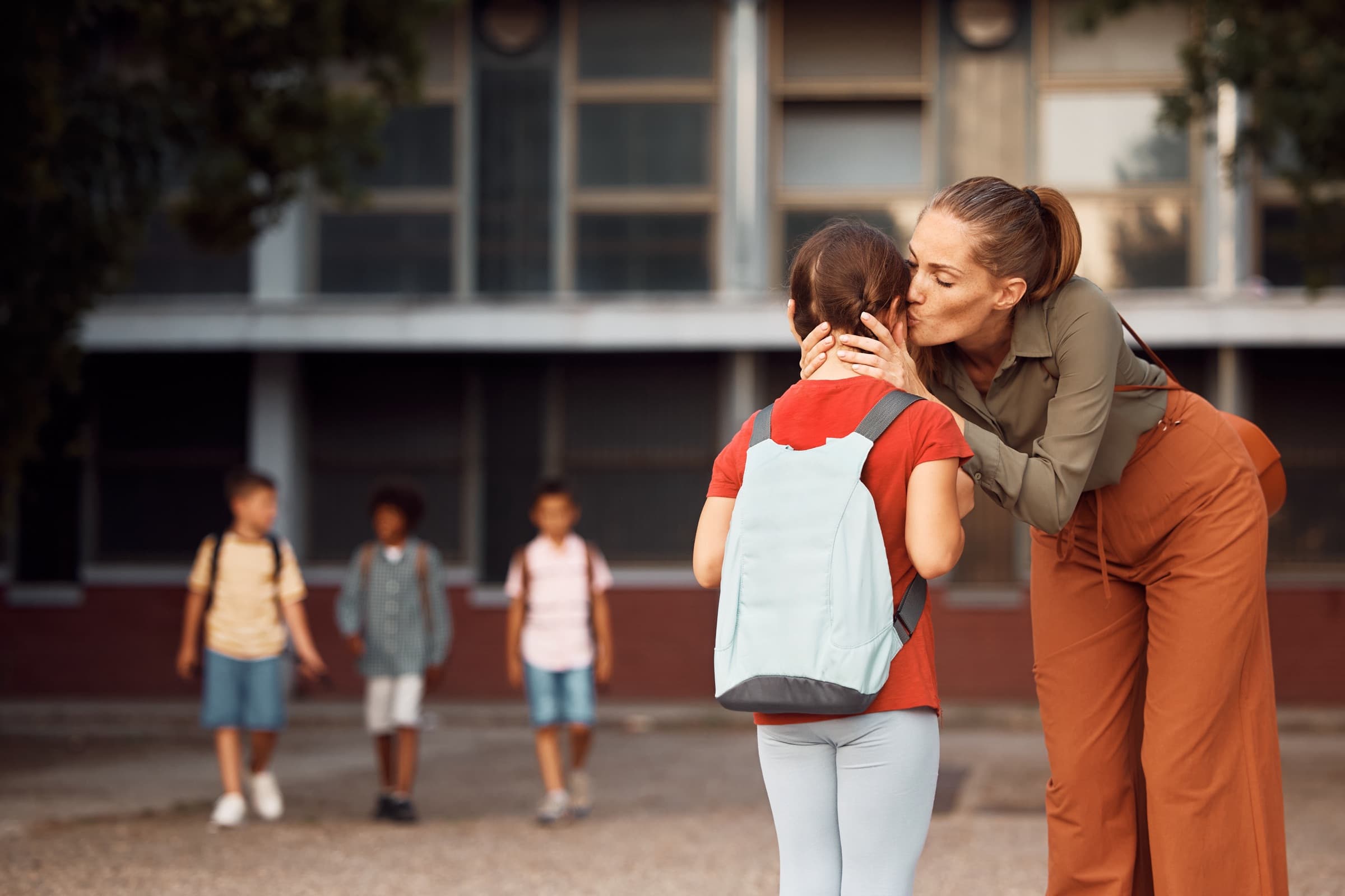 A mother kissing her daughter goodbye outside school as other children walk in