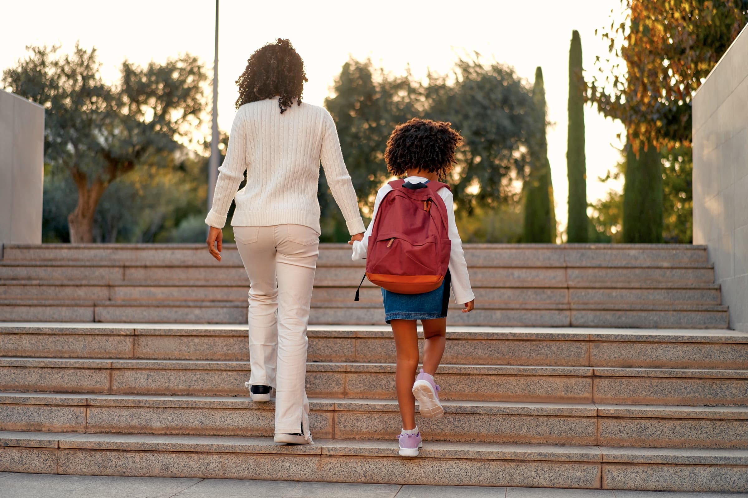 A mother and daughter with a backpack walking hand in hand up the steps to school