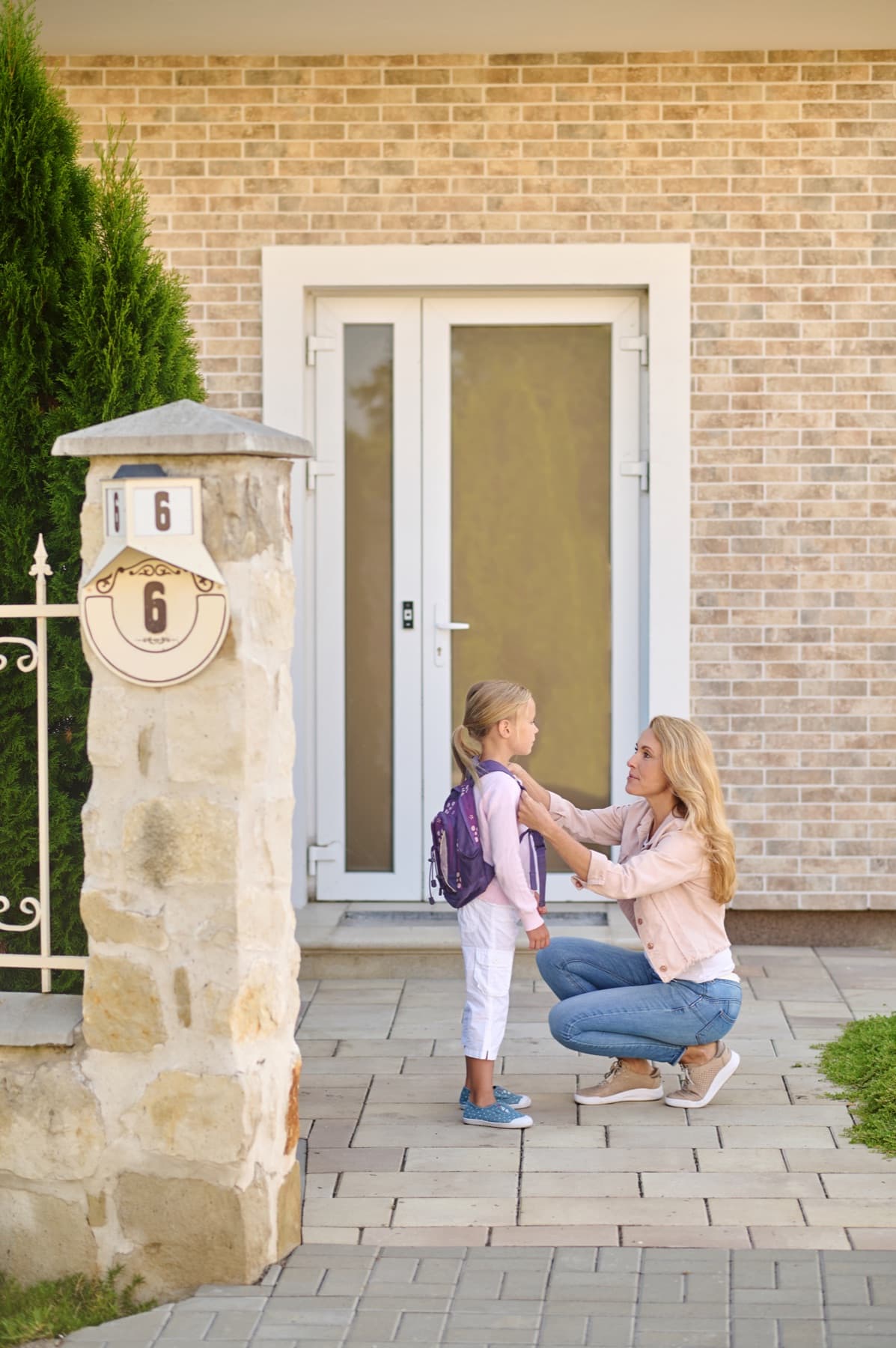 A mother kneeling to adjust her daughter's backpack outside the front door of their home